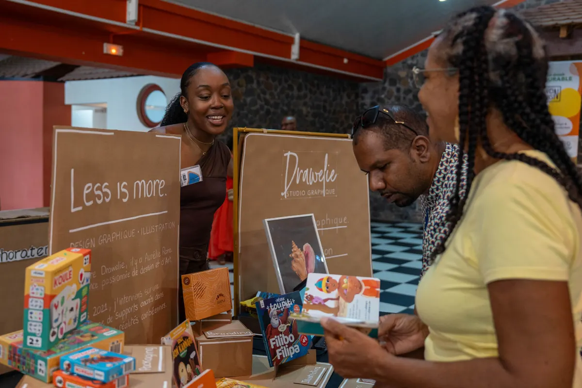Stand d'une designer graphique caribéenne au Salon du Design 2026, des visiteurs découvrent ses créations